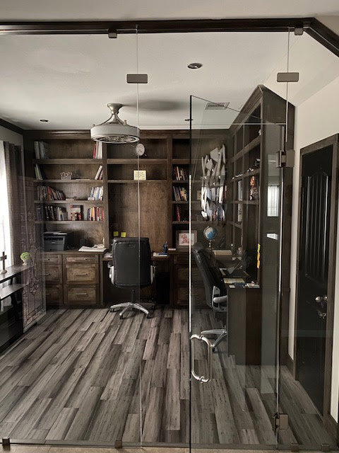 A custom home office with built-in wooden bookshelves and desks, featuring a glass partition, by MC Master Construction in League City, TX