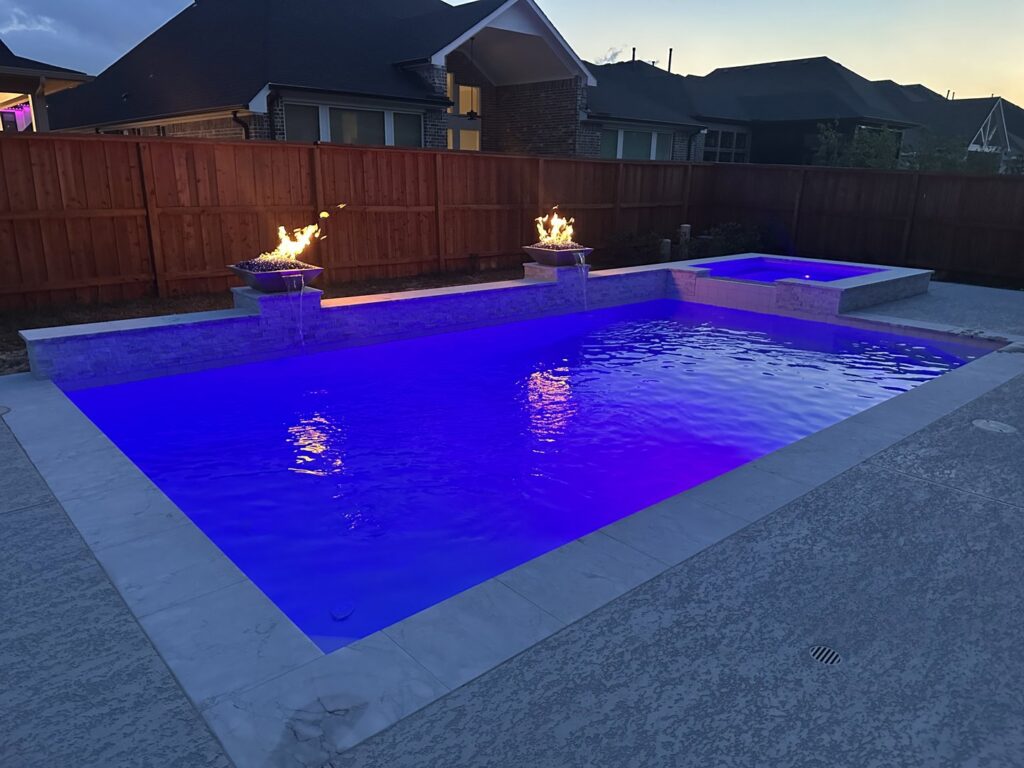 A custom pool with vibrant blue lighting, elegant fire bowls, and a relaxing spa at night by Limitless Custom Pools and Backyards in Houston, TX.
