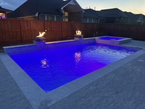 A custom pool with vibrant blue lighting, elegant fire bowls, and a relaxing spa at night by Limitless Custom Pools and Backyards in Houston, TX.
