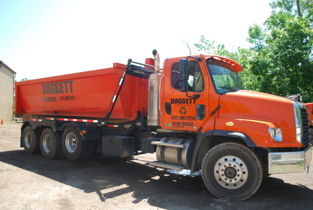 An orange Daggett Container Service LLC truck with a roll-off dumpster in bright daylight in Lansing, MI.