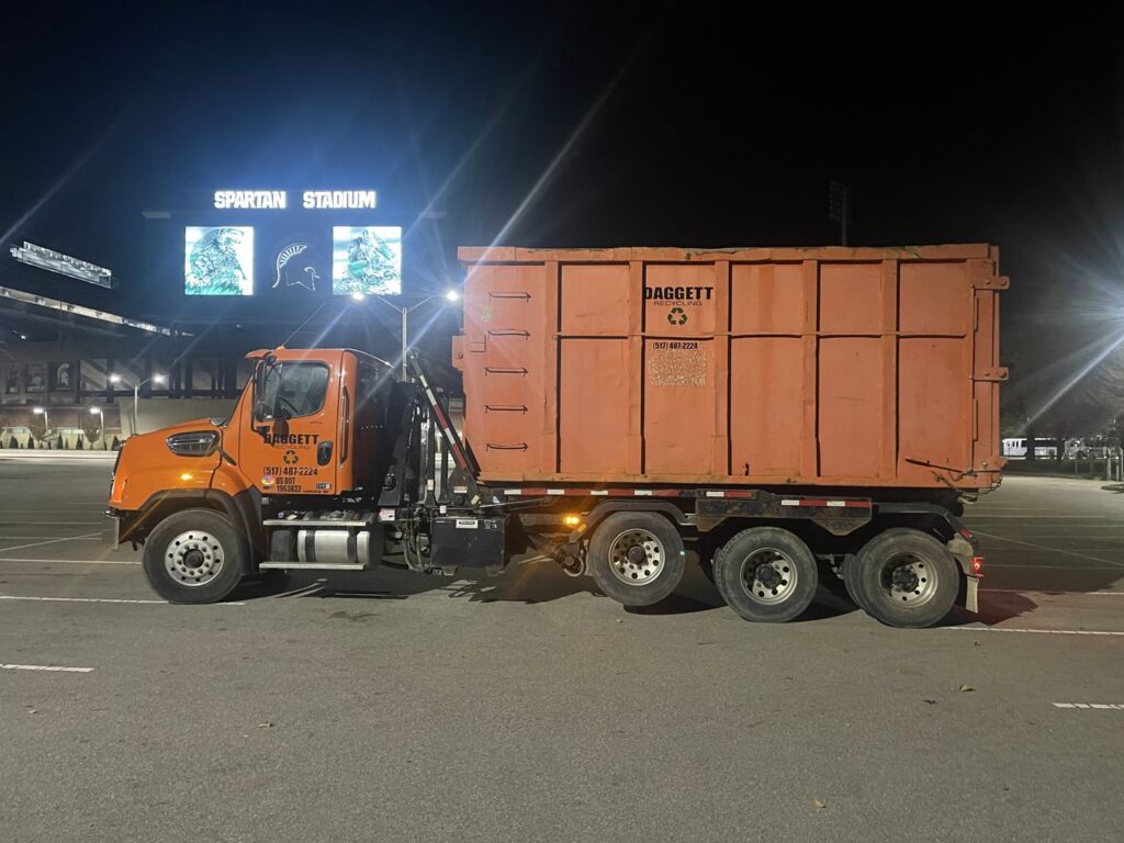An orange Daggett Container Service LLC truck with a dumpster parked at Spartan Stadium at night in Lansing, MI.