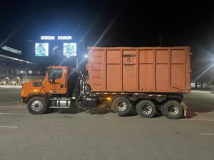 An orange Daggett Container Service LLC truck with a dumpster parked at Spartan Stadium at night in Lansing, MI.