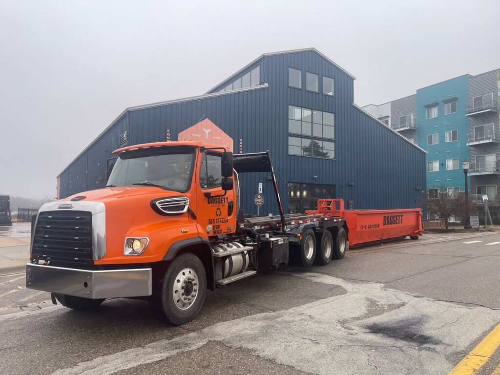 An orange Daggett Container Service LLC truck with a roll-off dumpster on a city street in Lansing, MI.