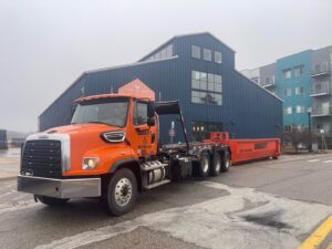 An orange Daggett Container Service LLC truck with a roll-off dumpster on a city street in Lansing, MI.