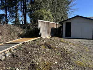 A damaged wooden fence section lying on the ground next to a shed, ready for repair by HandyMan Shocks in Tacoma, WA.