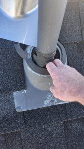 A hand pointing to a damaged roof vent boot on a shingle roof, indicating a repair needed by Elsass Home Services in Idaho Falls, ID.