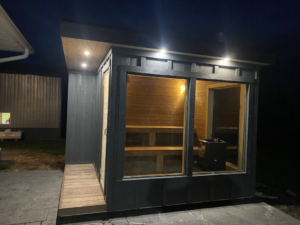 Exterior of a modern dark grey sauna at night with interior and exterior lights illuminated by No Boundaries Tiny Homes in Eau Claire, WI