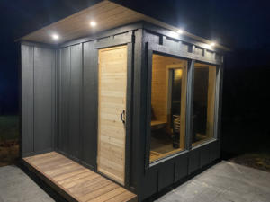 Exterior of a dark grey sauna at night with a wooden access ramp and illuminated interior by No Boundaries Tiny Homes in Eau Claire, WI