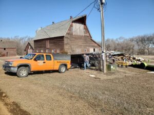 A BeeBop's Junk Removal truck and trailer parked near a large pile of wood debris next to an old barn in Sioux Falls, SD.