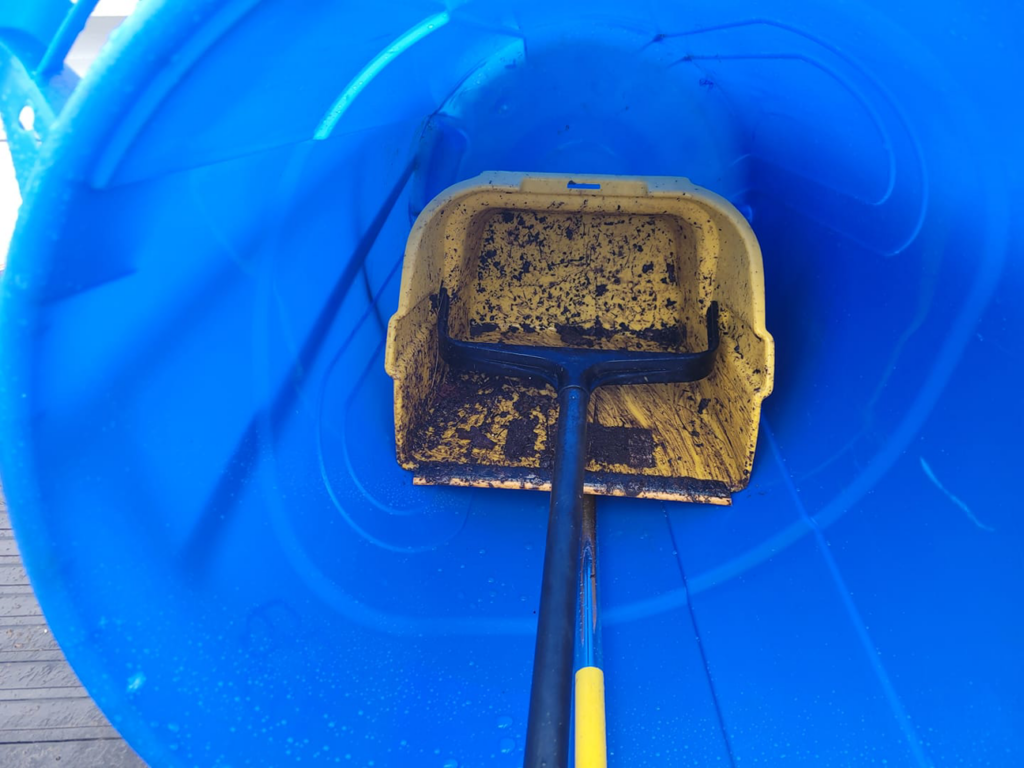 A yellow scoop with collected pet waste being emptied into a large blue disposal barrel by Slinging Scoops Co. in Chesapeake, VA.