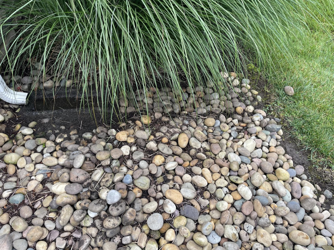 A downspout directing water into a river rock bed, showcasing exterior drainage solutions by AnderCall Professional Services LLC in Seaford, DE.