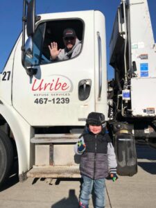 A Uribe Refuse Services driver waving from his garbage truck with a child standing nearby in Lincoln, NE.