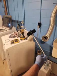 A handyman cleaning a dryer vent duct with a brush tool in a laundry room, a service provided by Cedar Valley Maintenance and Handyman Services LLC in Waterloo, IA.