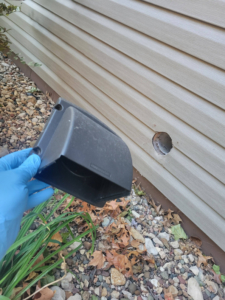 A gloved hand holding a dryer vent cover next to a hole in house siding, indicating dryer vent installation by Cedar Valley Maintenance and Handyman Services LLC in Waterloo, IA.