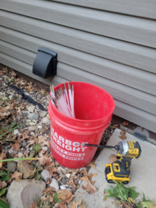 Dryer vent installed on a house exterior with cleaning tools and a drill on the ground, showing maintenance by Cedar Valley Maintenance and Handyman Services LLC in Waterloo, IA.