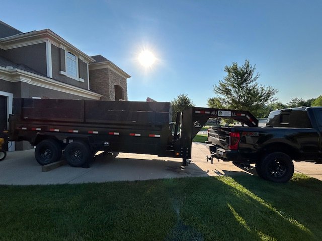 A black dump trailer attached to a pickup truck, parked in a residential driveway by D's Dumpster Rentals, Overland Park, KS.