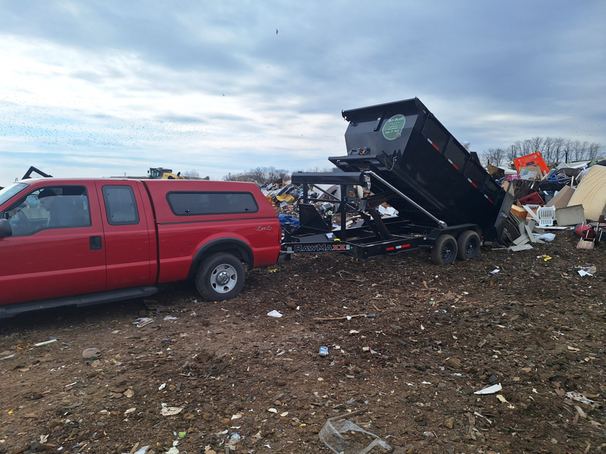 A red pickup truck towing a Bin Buds Dumpsters, LLC trailer, emptying a load of junk at a disposal site in Janesville, WI.