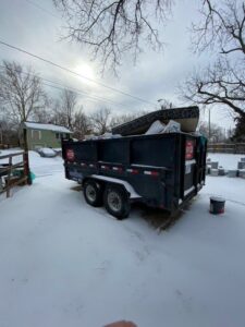 A dump trailer filled with junk, including a mattress, in a snowy residential area by D's Dumpster Rentals, Overland Park, KS.