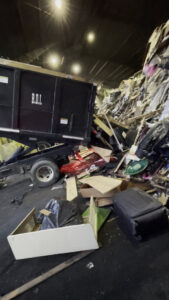 A black dump trailer being loaded with a large pile of mixed junk and debris inside a facility by Lemcor in Newark, NJ.