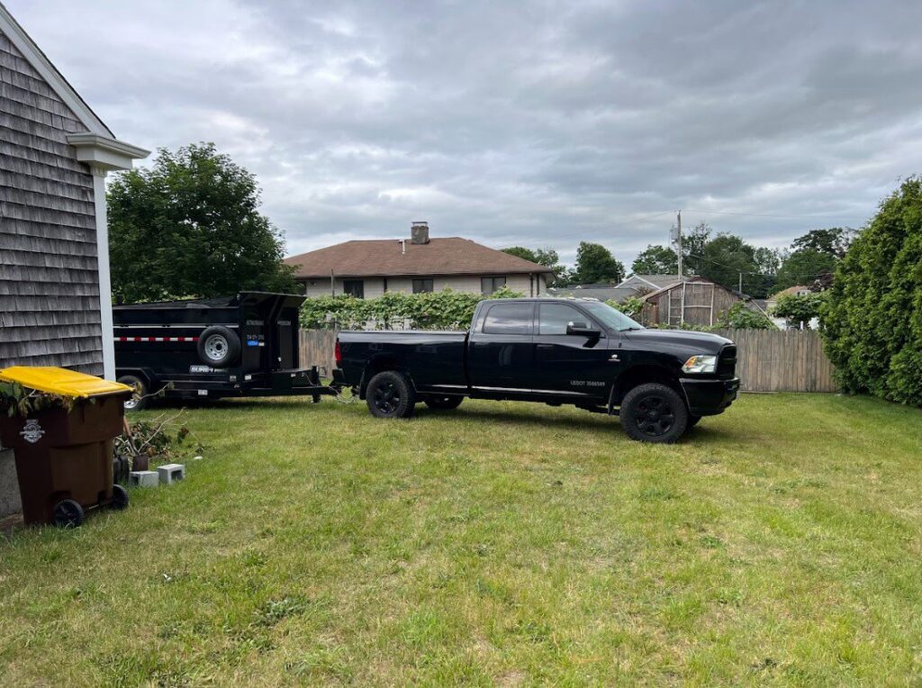 A black dump trailer parked on a residential lawn, ready for junk removal by Getting Trashed Dumpster Rentals & Excavation in Assonet, MA.
