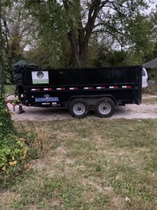 A black dump trailer with D's Dumpster Rentals logo parked on a grassy area in Overland Park, KS.