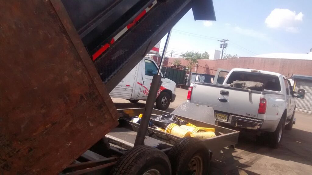 A dump trailer with its bed raised, showing yellow buckets and equipment, attached to a pickup truck for Lemcor in Newark, NJ.
