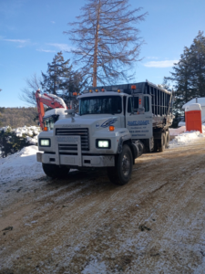 A dump truck and excavator on a snowy job site for East Coast Container Services LLC in Northwood, NH.
