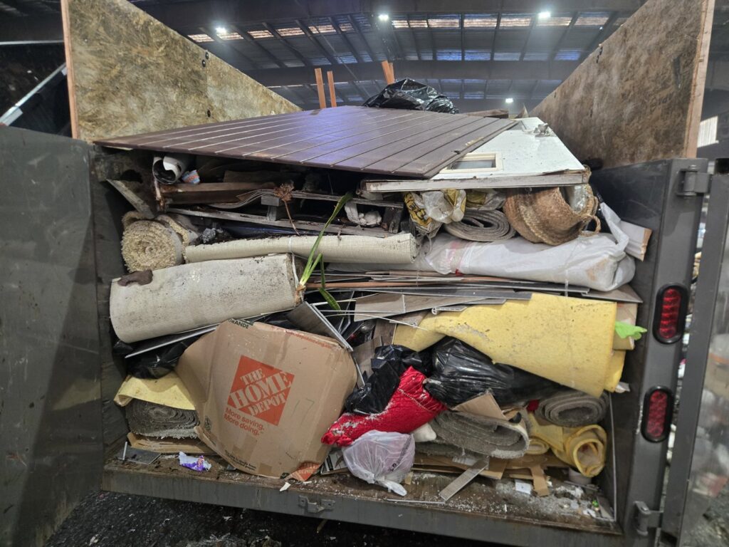 A large dump truck completely filled with various junk and debris, including carpets, wood, and boxes, by Titan Junk Co. in San Diego, CA