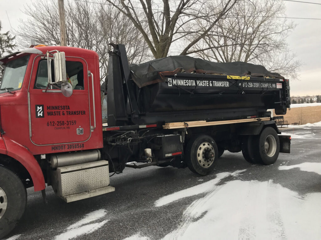 A red dump truck from Minnesota Waste and Transfer transporting a covered waste container on a snowy road in Champlin, MN
