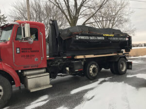A red dump truck from Minnesota Waste and Transfer transporting a covered waste container on a snowy road in Champlin, MN
