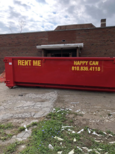 A red Happy Can Disposal dumpster parked in front of a brick building for junk removal services in Flint, MI.