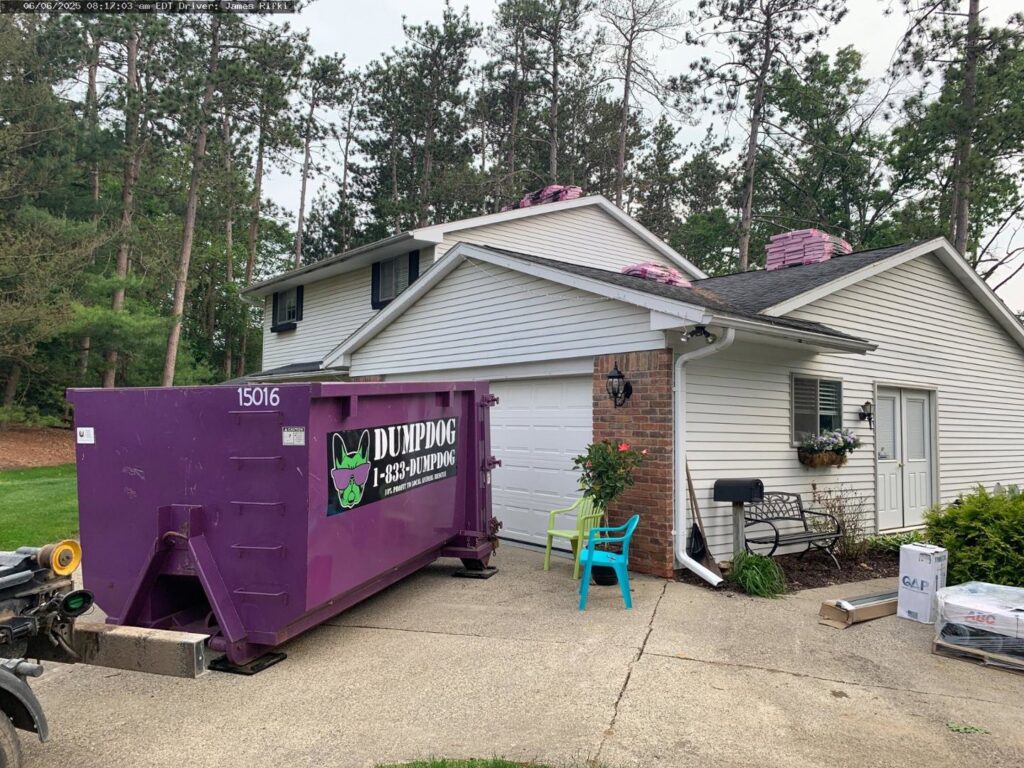 A DumpDog purple dumpster placed at a house undergoing renovation with roofing materials on the roof in Flint, MI.