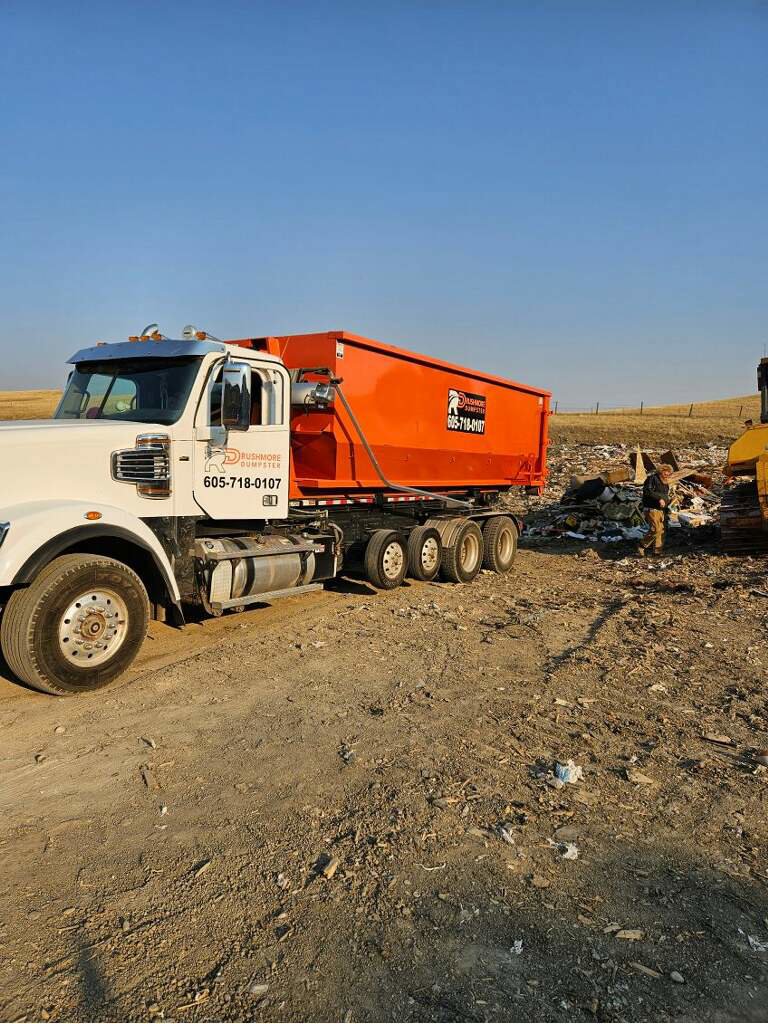 A Rushmore Dumpster truck with an orange roll-off dumpster at a landfill or waste transfer site in Rapid City, SD.