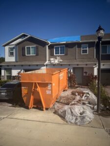 An orange dumpster placed at a residential home with roofing debris and a blue tarp on the roof by Pro Dumpsters & Junk Removal in Orlando, FL.