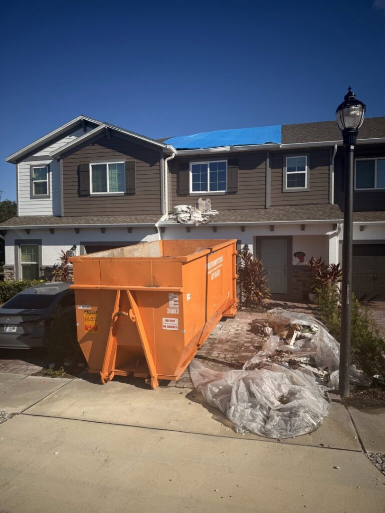 An orange dumpster placed at a residential home with roofing debris and a blue tarp on the roof by Pro Dumpsters & Junk Removal in Orlando, FL.