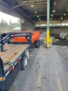A flatbed truck with an orange roll-off dumpster at a waste transfer station for ADE Hauling in Tacoma, WA