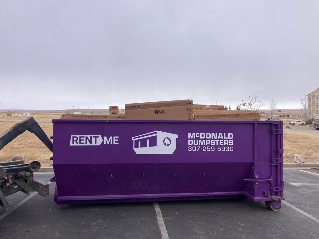 A McDonald Dumpsters purple dumpster filled with cardboard boxes being loaded onto a truck in Bar Nunn, WY.