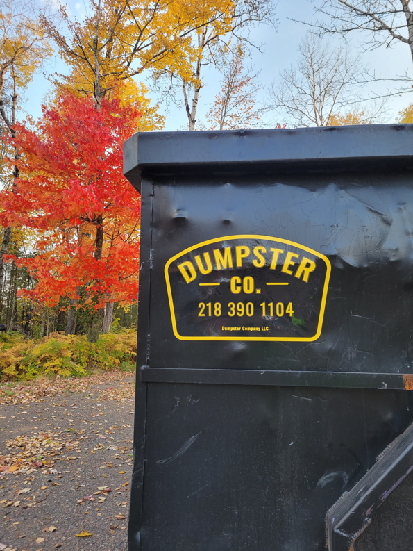 A black dumpster with the Dumpster Co logo, set against vibrant autumn trees in Duluth, MN.