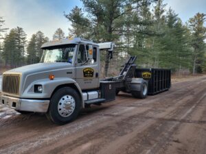 A Dumpster Co truck with a black dumpster parked on a dirt road in a natural setting, ready for junk removal in Duluth, MN.