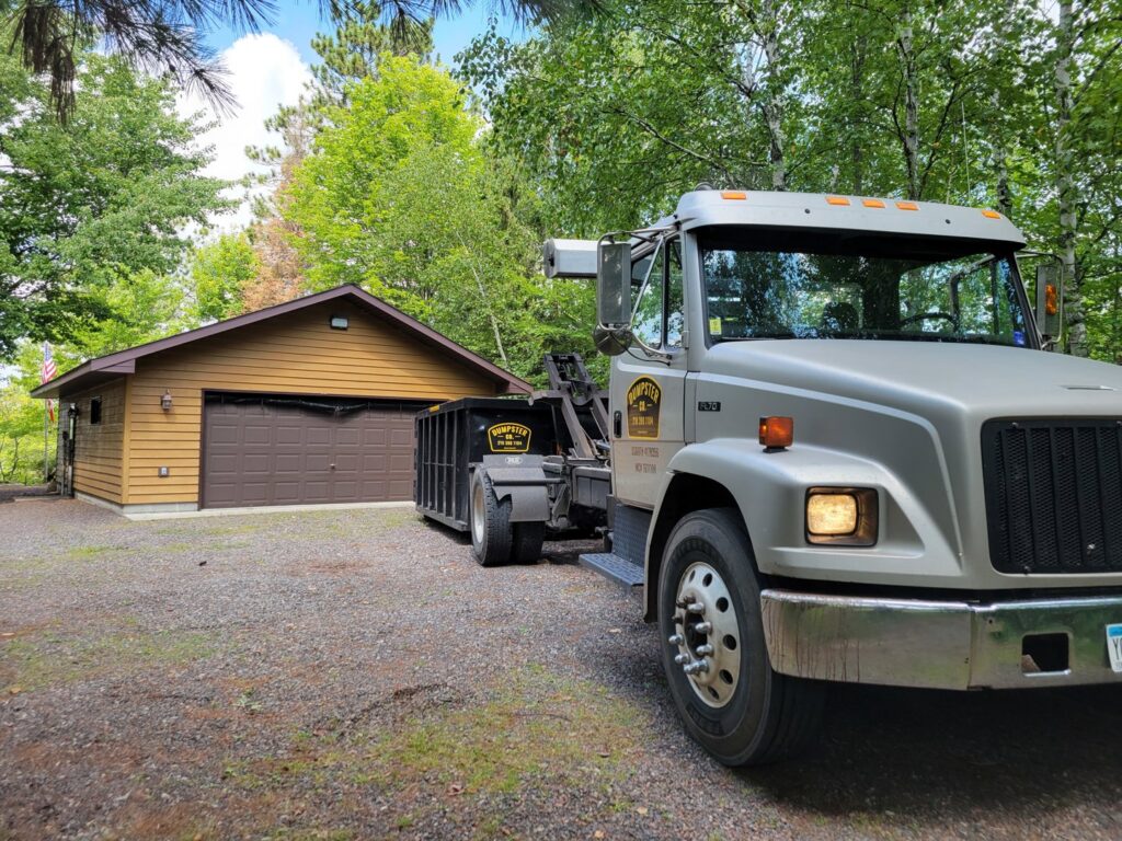 A Dumpster Co truck with a black dumpster parked in front of a residential garage, ready for junk removal in Duluth, MN.