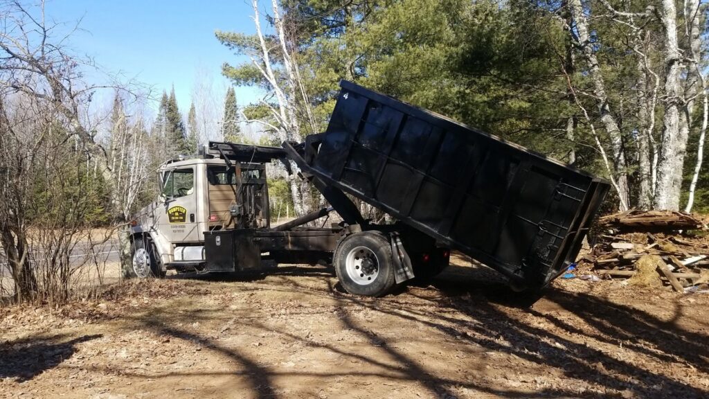 A Dumpster Co truck tilting a black dumpster in a wooded area, performing junk removal services in Duluth, MN.