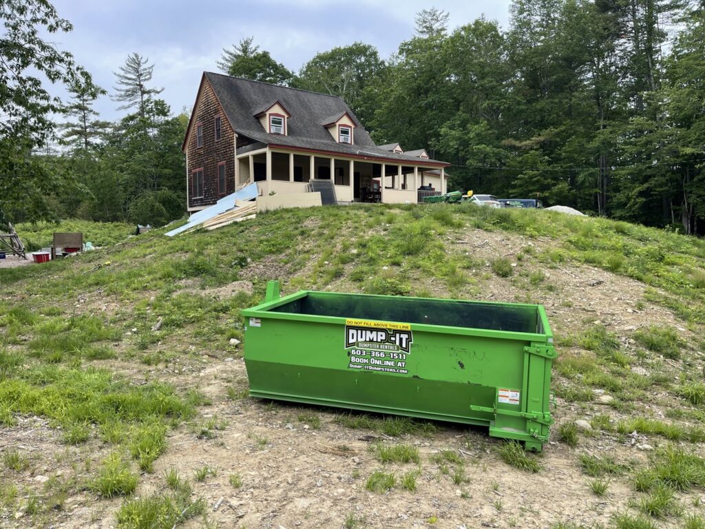 A green Dump-It Dumpster Rentals dumpster placed on a grassy hill at a construction or renovation site in Concord, NH.
