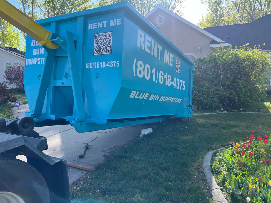 A blue dumpster being delivered by a truck to a residential driveway for Blue Bin Dumpster Rentals in Salt Lake City, UT.