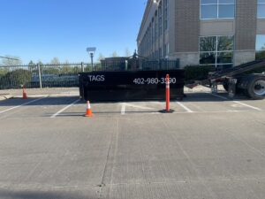 A black Tags Dumpsters roll-off dumpster placed in a parking lot with orange cones in Omaha, NE.