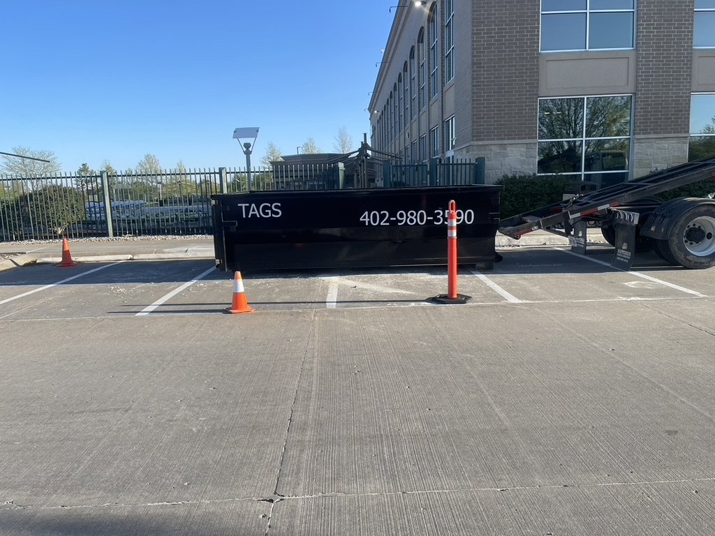A black Tags Dumpsters roll-off dumpster placed in a parking lot with orange cones in Omaha, NE.