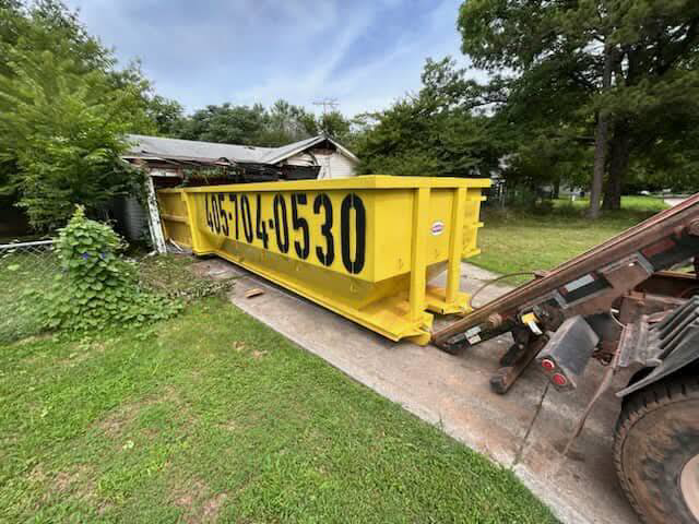 A yellow roll-off dumpster being delivered or picked up at a residential site by Tecumseh Rolloff Services in Moore, OK.