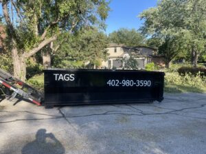 A Tags Dumpsters roll-off dumpster being delivered or picked up on a residential street with tree debris in Omaha, NE.