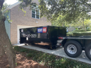 A black dumpster being delivered or picked up in a residential driveway by The Dumpster Dad in Wilmington, NC.