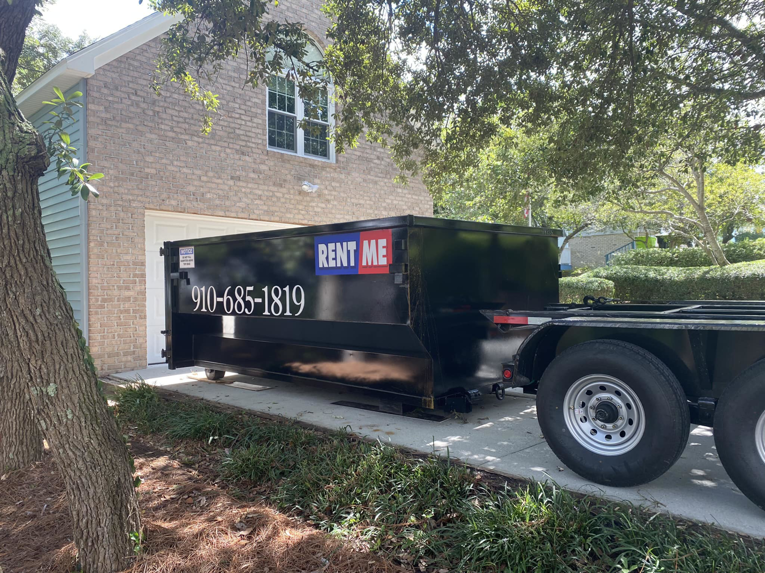 A black dumpster being delivered or picked up in a residential driveway by The Dumpster Dad in Wilmington, NC.