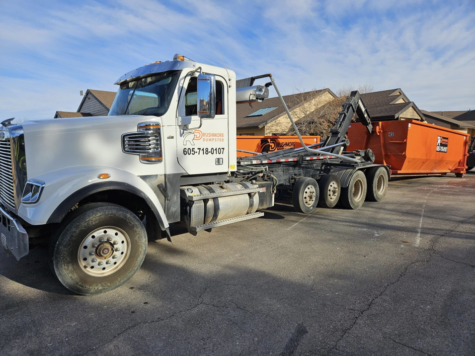 A Rushmore Dumpster truck delivering an orange roll-off dumpster to a residential area in Rapid City, SD.